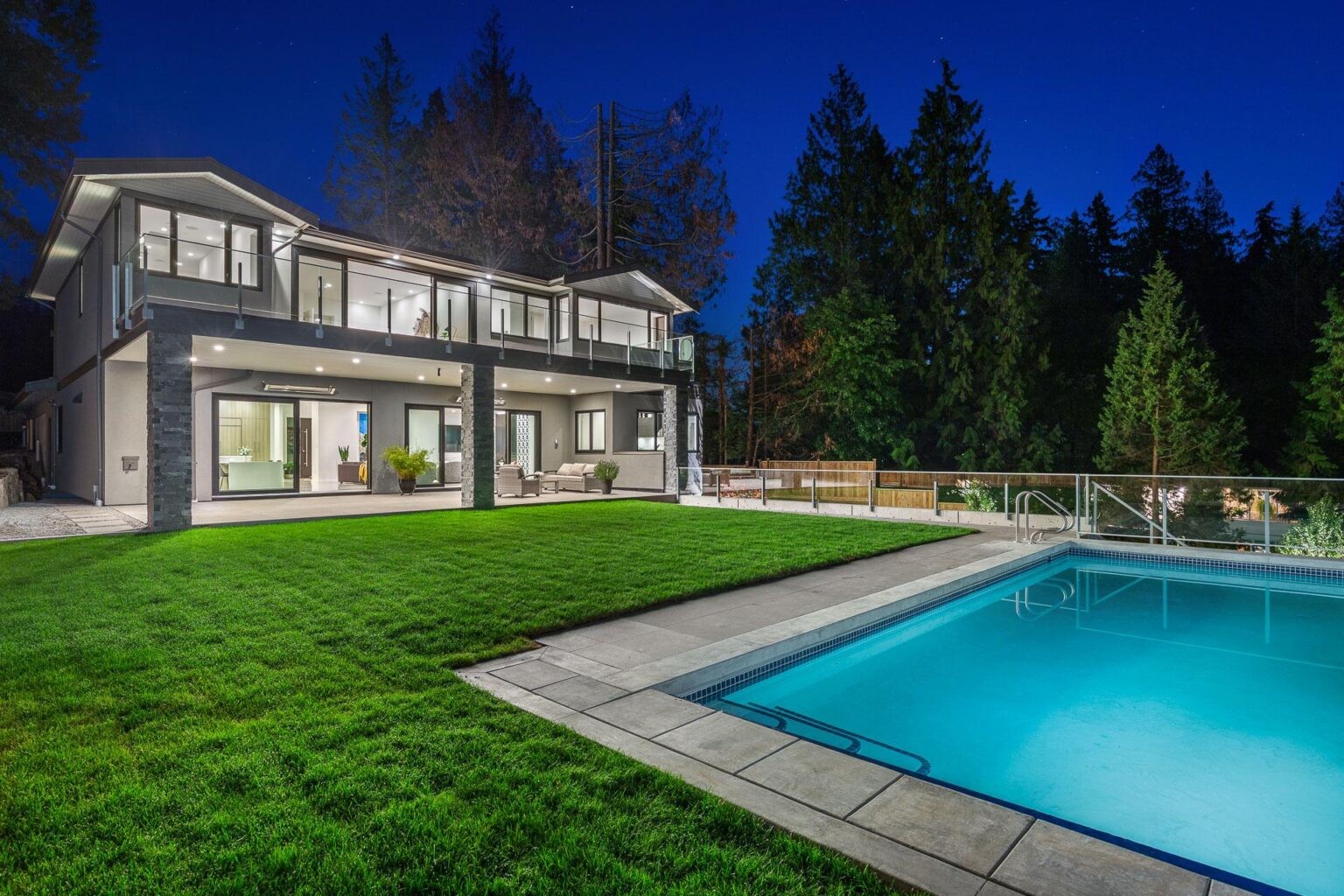 Modern luxury home with glass balcony, illuminated exterior, landscaped lawn, and swimming pool at night.